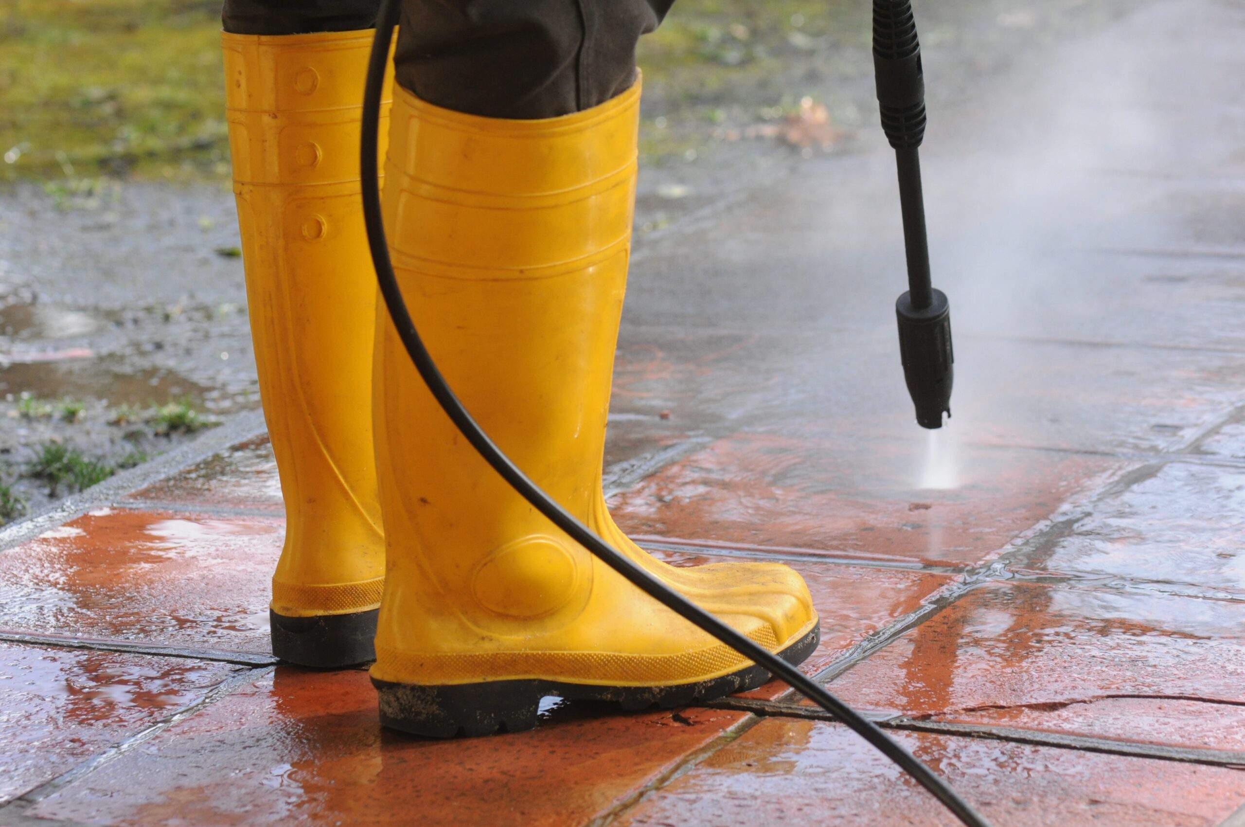 person wearing yellow rubber boots with high pressure water nozzle cleaning dirt tiles scaled TheHomeQuality